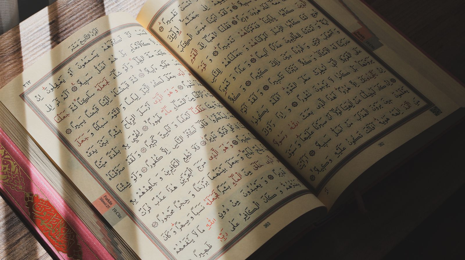 مصحف مفتوح على صفحتين من سورة الفرقان، تُضيئه أشعة شمس مائلة فوق طاولة خشبية.
