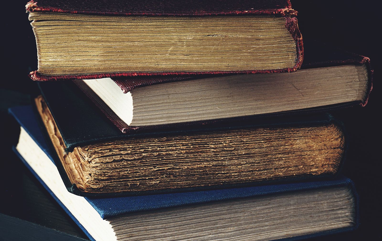 تكديس عن قرب لكتب قديمة بأغلفة جلدية وحواف صفحات مذهّبة باهتة في إضاءة داكنة هادئة