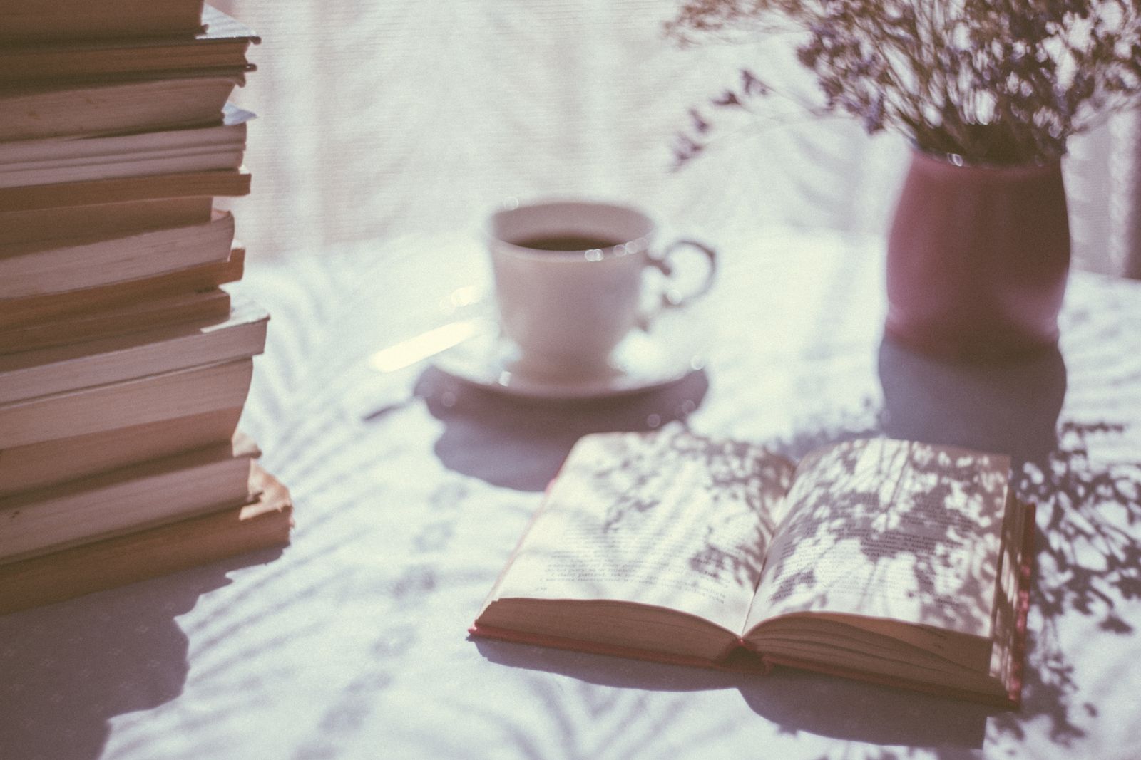 كتاب مفتوح بجانب فنجان قهوة وإناء زهور صغير على طاولة مضاءة بنور دافئ متداخل الظلال، بلا أي أشخاص ظاهرين في المشهد