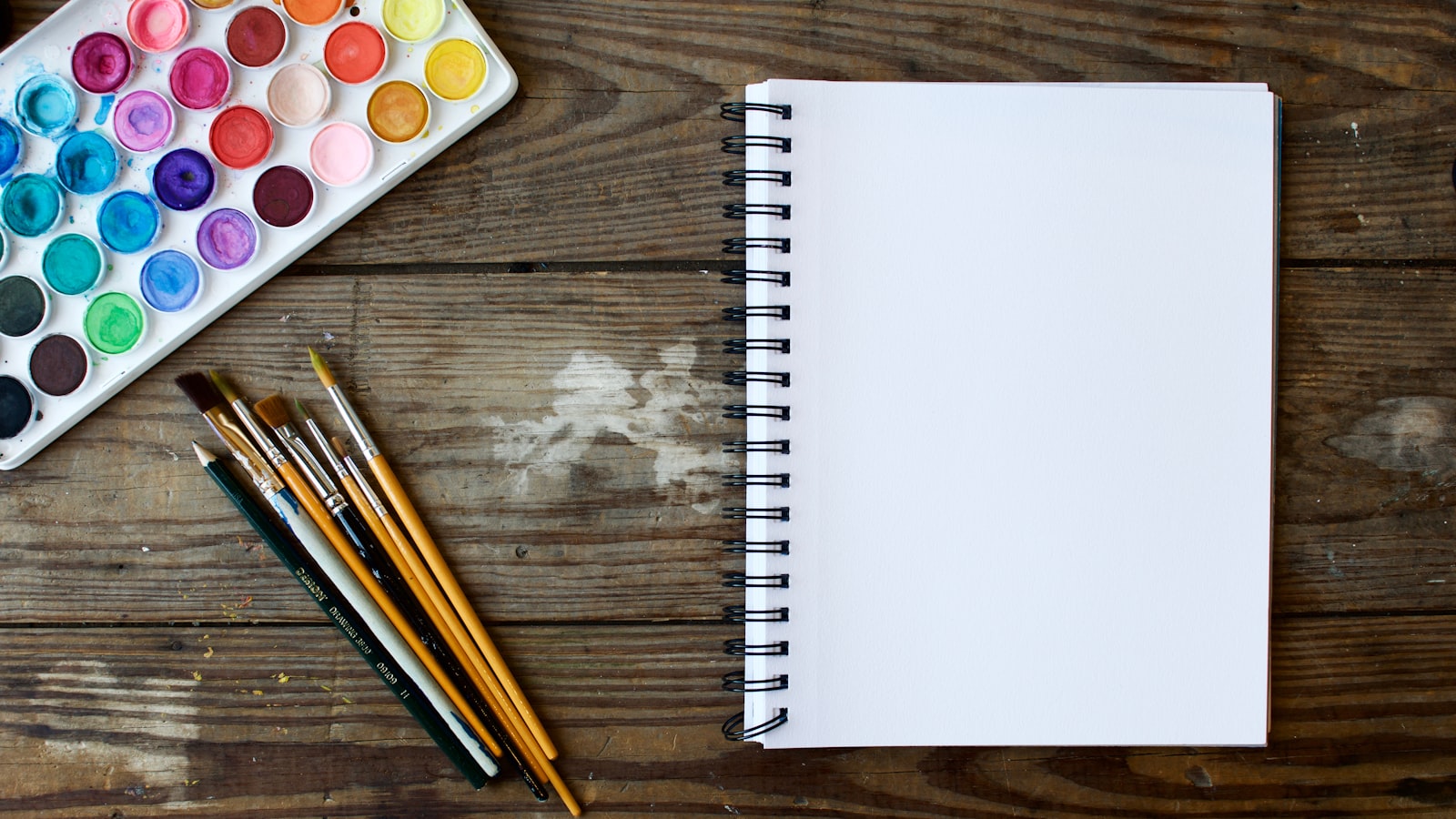 فرشاة رسم وباليت ألوان مائية بجانب دفتر رسم أبيض فارغ على طاولة خشبية